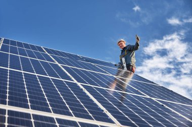 Neşeli erkek işçi güzel bulutlu gökyüzü altında mavi fotovoltaik güneş paneli kurarken başparmak sallıyor ve gülümsüyor. Elektrikçi onay jesti gösteriyor. Alternatif enerji kavramı