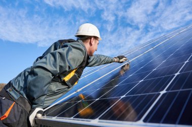 Side view snapshot of workman, wearing uniform, working gloves and helmet, setting a shiny new solar battery with help of hex key, blue sky on background. Green energy concept clipart