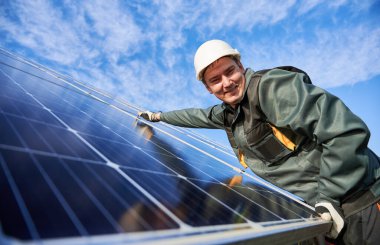 Mavi fotovoltaik güneş paneli yerleştirirken kameraya gülümseyen neşeli erkek işçi. Güzel gökyüzünün altında kasklı bir elektrikçi. Alternatif enerji kaynakları ve yenilikler kavramı.