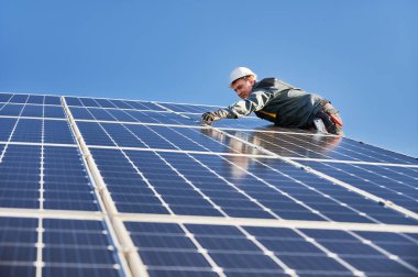 Erkek işçilerin güneş modülleri kurarken ve fotovoltaik güneş dizisini desteklerken yan görünümü. Güneş paneli ile çalışırken güvenlik kaskı ve eldiven takan bir elektrikçi. Güneş enerjisi kavramı