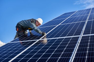 Güzel ve parlak güneş pili yüzeyi ve merdivende duran üniformalı bir işçi. Erkek elektrikçi, güneşli bir günde güneş modülleri kuruyor. Düşük açılı. Alternatif enerji kavramı