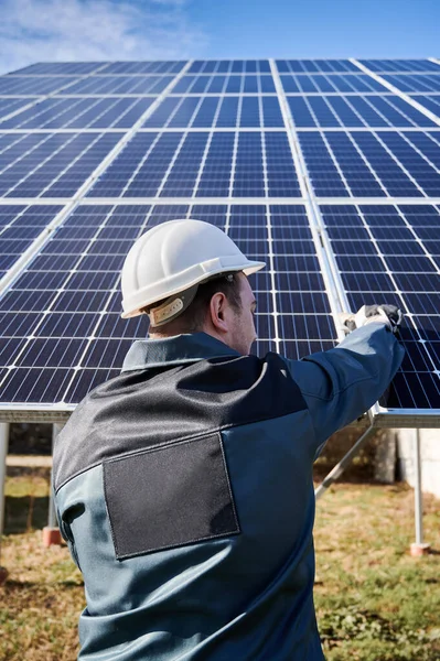 Back view of man electrician in safety helmet mounting blue photovoltaic solar panel. Solar installer mounting solar modules of solar array. Maintenance concept