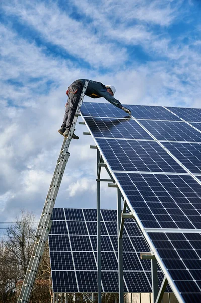 Vertical shot of man worker electrician repairing photovoltaic solar module, standing on ladder under beautiful cloudy sky, installing solar photovoltaic panel. Concept of alternative energy