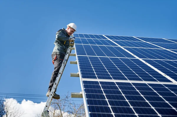Electrician on ladder installing photovoltaic solar panel under blue sky. Man technician wearing safety helmet and gloves while mounting solar module. Concept of sun energy, solar panel installation.
