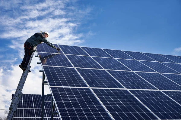 Male worker in white safety helmet standing on ladder and repairing photovoltaic solar panel station under beautiful blue cloudy sky. Concept of alternative energy and power sustainable resources.