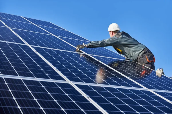 Worker standing on ladder and installing photovoltaic solar panel system. Male electrician in white safety helmet under blue sky. Concept of alternative energy and power sustainable resources.