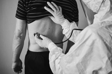PPE koruyucu kıyafetli bir doktorun yakın plan fotoğrafı. Evde yaşlı bir erkek hastayı fonendoskop kullanarak muayene ediyor. Sırtını kontrol ediyor, siyah beyaz.