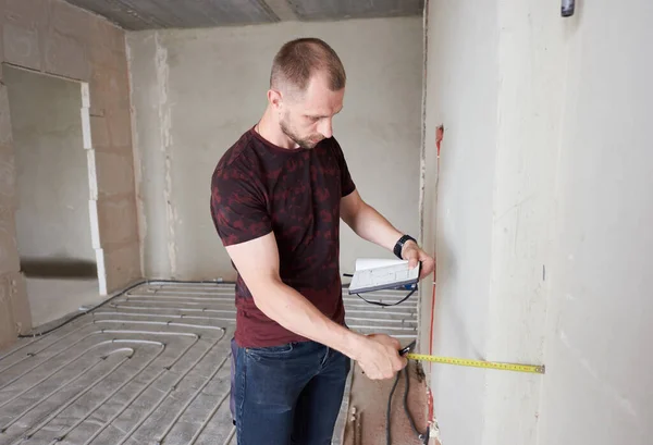 Side view of serious man in shirt holding notebook and using measuring tape in kitchen with underfloor heating pipes. Handyman taking measurement while working on renovation of apartment.