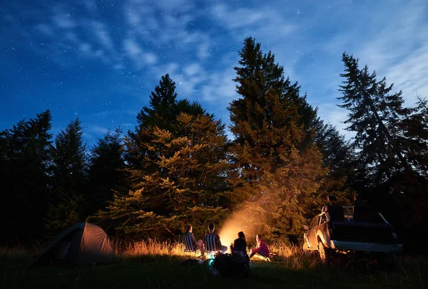 Night starry sky over meadow with travelers near campfire. Tourists resting in the wood with tall coniferous trees, car and camp tent under blue sky. Concept of travelling and night camping.