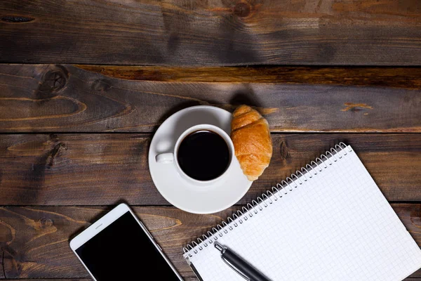 Weiße Tasse Kaffee und Handy und neben Notizbuch mit Stift, Croissant liegen — Stockfoto