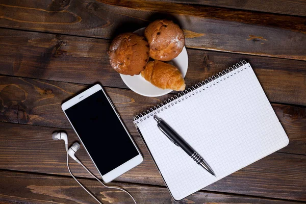 Белая чашка кофе с наушниками и мобильным телефоном и лежащая рядом с ноутбуком с ручкой, круассаном — стоковое фото