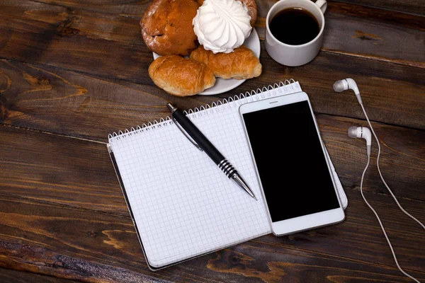 Белая чашка кофе с наушниками и мобильным телефоном, лежащая рядом с ноутбуком с ручкой, круассаном, зефиром — стоковое фото