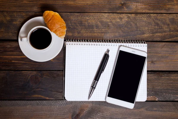 Vista de cima. Telefone celular com xícara branca de café e bloco de notas em fundo de madeira. Espaço de trabalho flat lay . — Fotografia de Stock