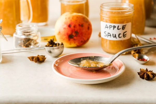 Applesauce buatan sendiri yang baru dimasak dalam toples kaca, bahan-bahan, piring merah muda dan sendok di atas meja dapur, tampilan closeup — Stok Foto