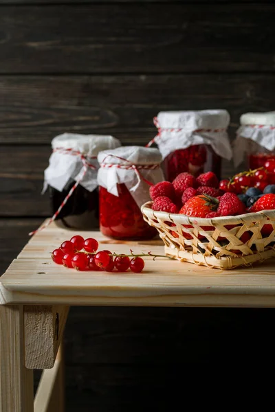 Berbagai macam buah beri dalam keranjang, toples kaca dengan selai dari buah-buahan yang berbeda di atas meja kayu . — Stok Foto