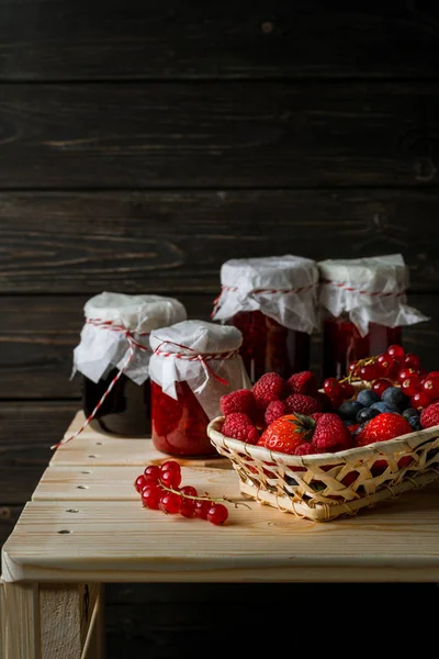 Berbagai macam buah beri dalam keranjang, toples kaca dengan selai dari buah-buahan yang berbeda di atas meja kayu . — Stok Foto