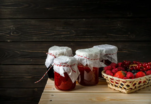 Натюрморт з певними ягодами в кошику, скляні банки з джемами з різних ягід на дерев'яному столі . — стокове фото