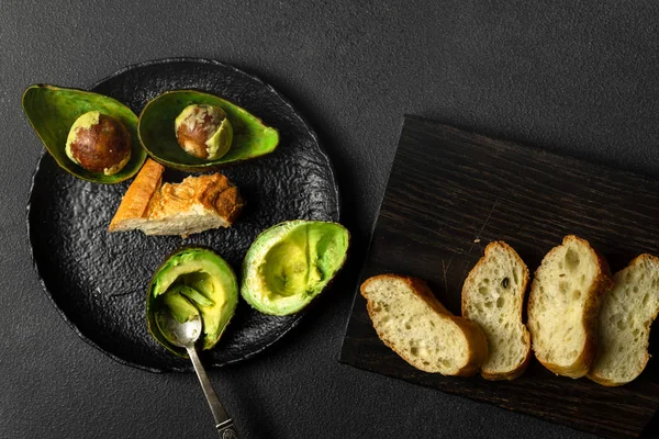 Avocado halves, spoon on black plate, bread on black board on dark background, top view — 스톡 사진