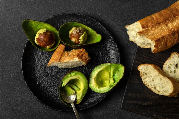 Mitades de aguacate, cuchara sobre plato negro, pan sobre pizarra negra sobre fondo oscuro, vista superior — Foto de Stock