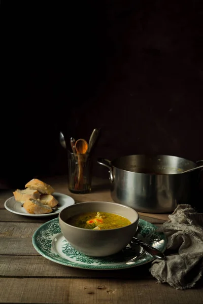 Natureza morta com sopa caseira de macarrão de frango, panela de cozinhar, pão, talheres em um copo em fundo rústico de madeira — Fotografia de Stock