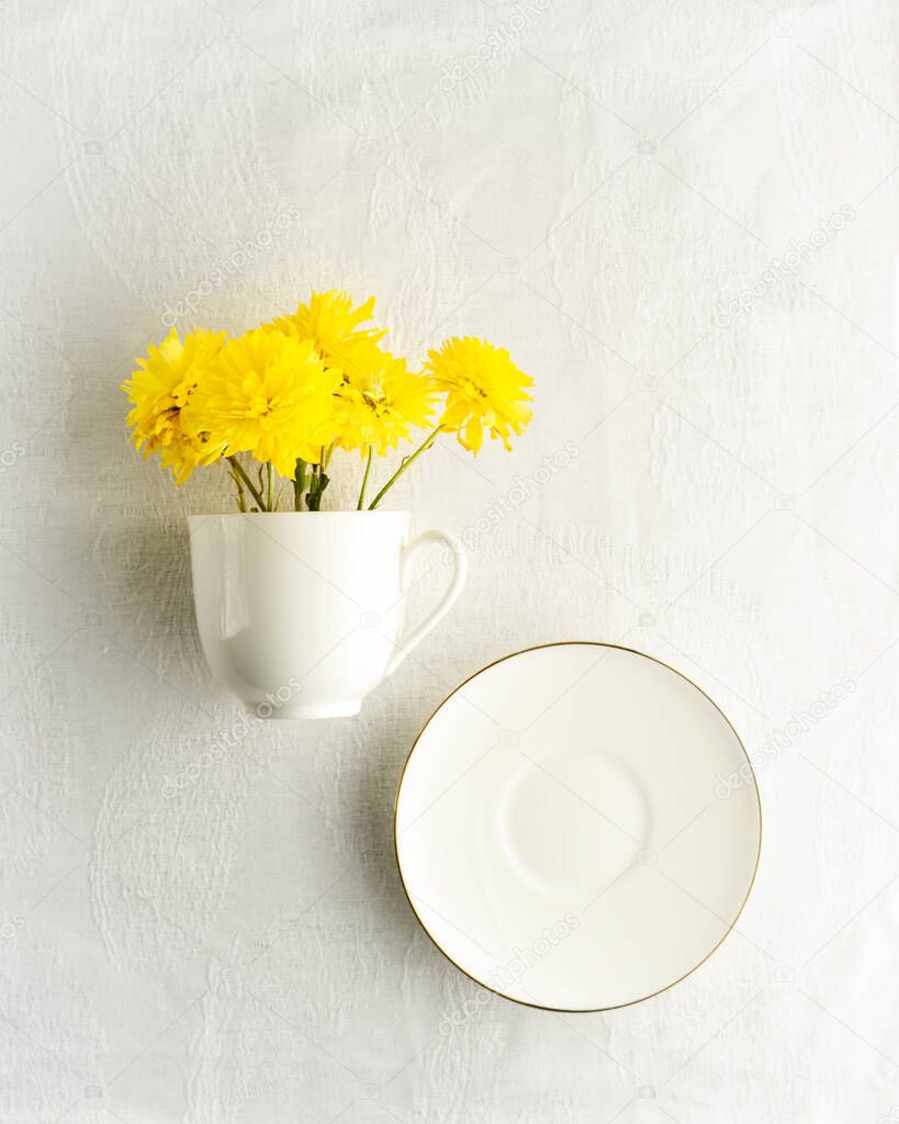 Small bouquet of yellow spring flowers in white cup, saucer on white tablecloth, top view