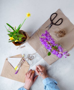 Günlük giysiler içindeki kadın hediye olarak kahverengi kağıda çiçek paketliyor, kartpostal yazıyor. Üst görünüm, dikey görüntü