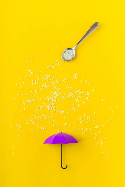 Rice grains pouring from spoon on purple toy umbrella on yellow background. Artistic concept of spring rain. Portrait orientation with copy space