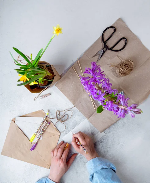 Woman in casual clothes packing spring potted flowers in brown paper as gifts, writing poscards. Top view, verical image