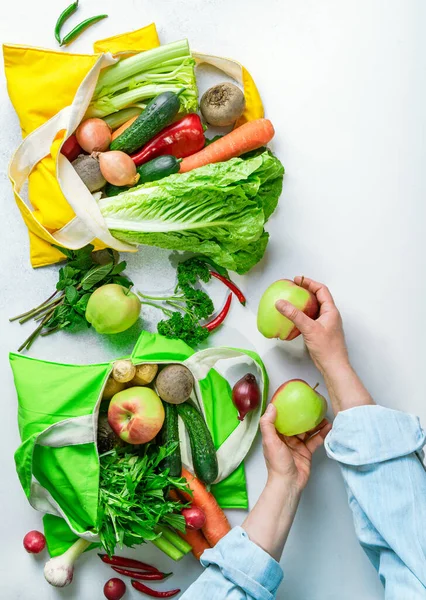 Textile shopping bags full of colorful vegetables and fruit on white background, woman\'s hands unpacking bags. Healthy diet or spring detox concept. Vertical image