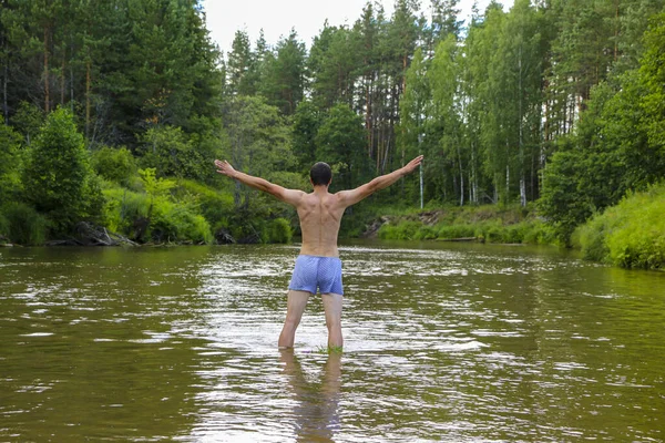 a young slender brunette man with his back standing in the water of a fast shallow river surrounded by a dense green forest. relaxation and unity with nature in isolation from people.