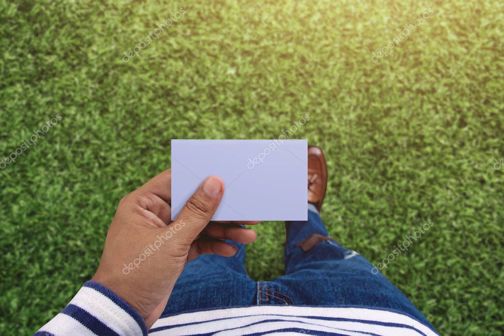 Blank business card on hand for mock up, Top view, present by casual urban guy stand over green grass field background