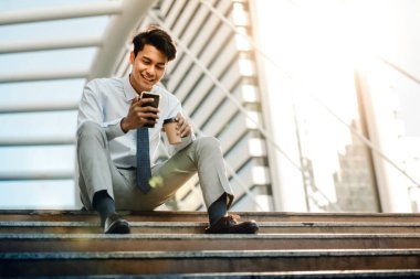 Happy Young Businessman Sitting on Staircase and Using Smartphone. Urban Lifestyle. Low Angle View. Modern Buiding in City as background