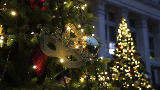 クリスマスツリーの背景とクリスマスの装飾 夕方に緑のモミにカラフルなボールと黄金の仮面舞踏会のマスク きらめくガーランドライトぼやけた背景 — ストック動画