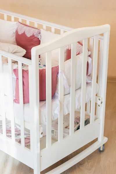 Baby bed with white and Burgundy color pillows with laces. Cot on wheels in the bedroom with pastel yellow colors in the morning. Harsh sun light come from window.Minimalistic interior