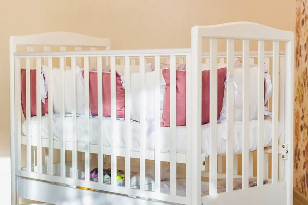 Baby bed with white and Burgundy color pillows with laces. Cot on wheels in the bedroom with pastel yellow colors in the morning. Harsh sun light come from window.Minimalistic interior