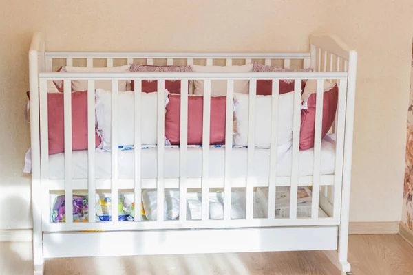 Baby bed with white and Burgundy color pillows with laces. Cot on wheels in the bedroom with pastel yellow colors in the morning. Harsh sun light come from window.Minimalistic interior