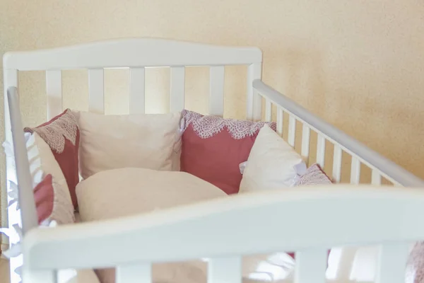 Inside baby bed with white and Burgundy color pillows with laces. Cot on wheels in the bedroom with pastel yellow colors. Minimalistic interior