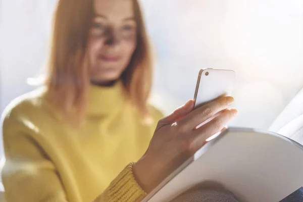 Young hipster girl checking new messages while reading magazine at home