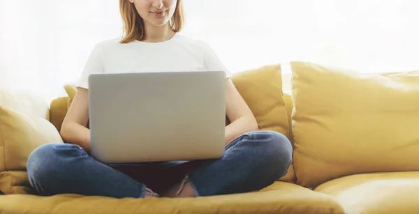 흰색 t-셔츠 그리고 청바지 편안한 소파에 앉아있는 동안 노트북을 사용 하 여 착용 하는 여자를 웃 고 — 스톡 사진