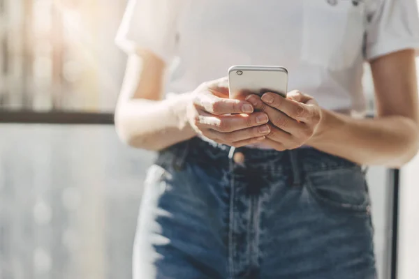 Hipster flicka klädd i vit t-shirt och blå jeans att skriva på pekskärm av mobiltelefon, närbild kvinnliga händer med moderna smartphone i city på bakgrunden — Stockfoto