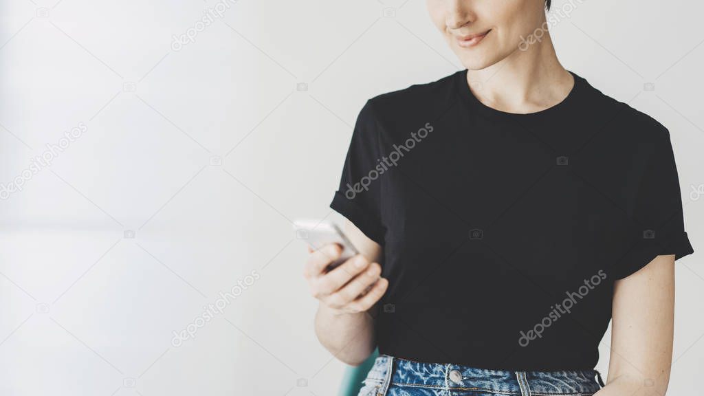 Mock-up of black cotton t-shirt, Young hipster girl wearing black t-shirt and using modern smartphone with white wall in the background