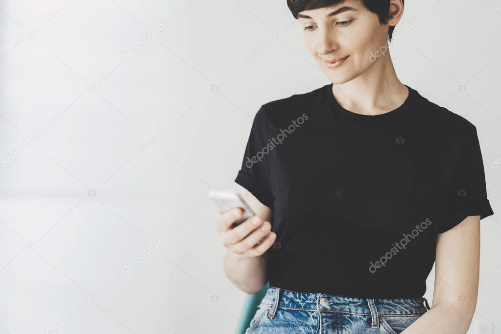 Portrait of young woman using modern smartphone with white wall in the background, Mock-up of black cotton t-shirt