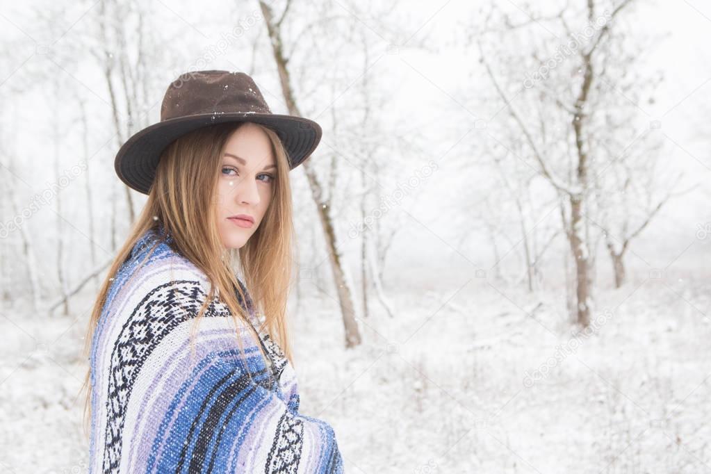 Young woman standing in snow with a bohemian hat and blanket near trees. 