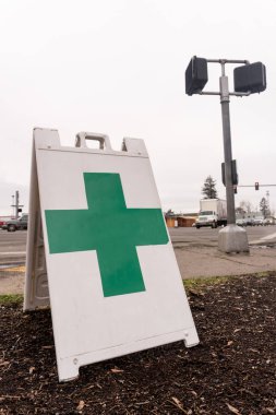 A free standing sign with a green cross shows that a marijuana dispensary is nearby high traffic busy intersection. clipart