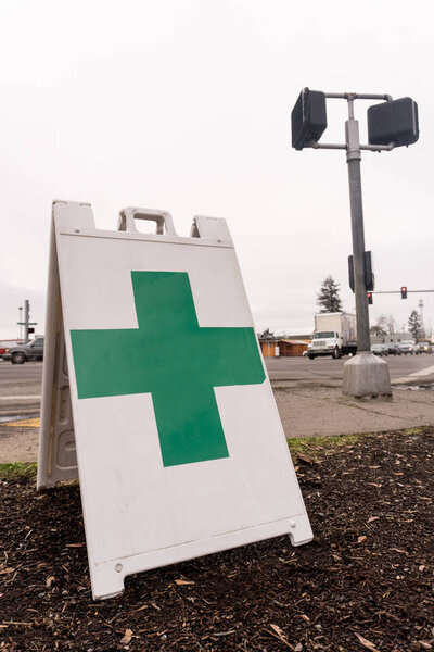 A free standing sign with a green cross shows that a marijuana dispensary is nearby high traffic busy intersection.