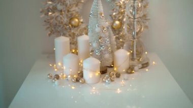 Beyaz ve altın renkli Noel süslemeleri, arka planda parlayan bokeh ışıkları. Küçük topları olan güzel bir yeni yıl ağacı. Beyaz mumlar ve sarı çelenklerle dolu bir ev