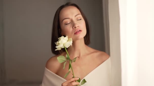 白のインテリアで白いバラと美しい女性のクローズアップ肖像画 ウィンドウで若い魅力的な自信のブルネットの女性 女性の自然の美しさ スローモーションビデオ — ストック動画