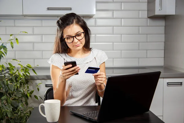 Jong meisje maakt aankopen online met credit card en telefoon zit aan de keukentafel — Stockfoto