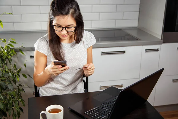 Молодая женщина делает онлайн покупку с помощью кредитной карты и телефона, сидящего на кухне — стоковое фото