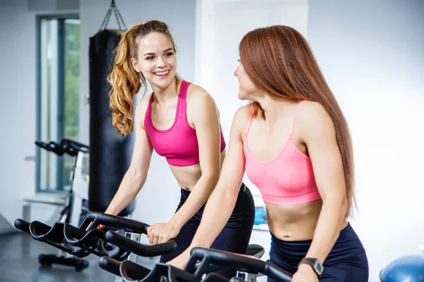 Dos mujeres bonitas haciendo ejercicios en bicicletas en el gimnasio — Foto de Stock